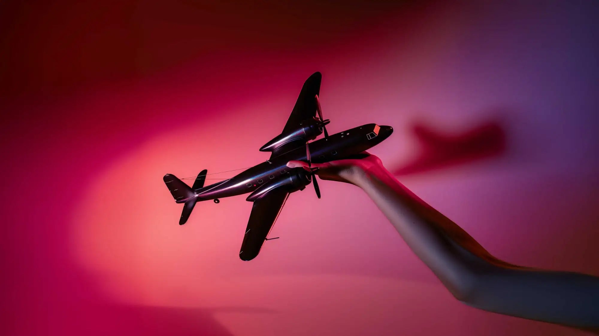 Un elegante avión modelo negro con un acabado brillante, sostenido suavemente por una mano contra un fondo vibrante de degradado rojo y púrpura.